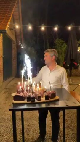 A man and a woman are celebrating a birthday outside at night. The woman, with blonde hair, is on the left in a floral print top and dark pants. She initially holds something blue in her hands, possibly a gift. The man is on the right wearing a light button-down shirt and dark pants. They stand beside a black table laden with pieces of chocolate cake. Each cake piece has a candle lit on top, and some also have sparklers going off. Behind them, strung overhead, are strings of lights. The scene is lit by these lights, candles, and sparklers. To their left, a brick building with a red-tiled roof and a dark green door can be seen. An open umbrella is behind the man, to his right. The man has his arm around the woman’s shoulder and kisses her head.