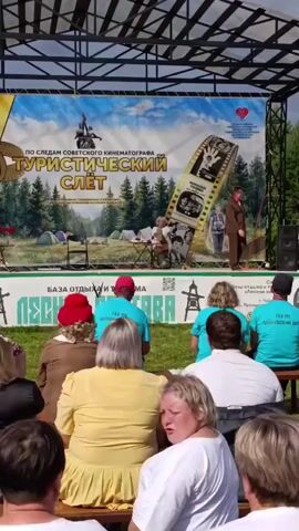 An outdoor stage is set up with a large banner in the background depicting a landscape with trees and tents, and text that appears to read "TOURIST CAMP" in large letters. On the stage, a man in a suit is walking from right to left, while a woman sits in a chair and appears to be speaking or performing. Several people are seated in the audience in the foreground, facing the stage. The environment appears to be a grassy area with trees visible in the background.