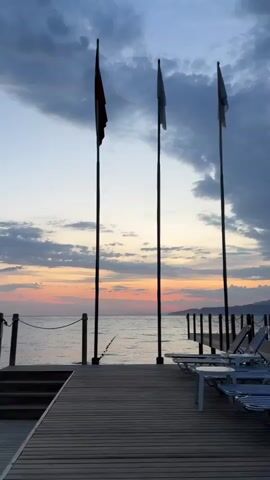 The video captures a serene sunset view from a wooden pier extending over a calm body of water. In the foreground, several blue and white striped lounge chairs with small white side tables are neatly arranged on the pier, facing the water. Four tall flagpoles with flags (one distinctly red, the others dark or black) stand prominently on the pier.

As the camera pans slightly to the right, a dark canopy structure becomes visible on the pier. In the distance, a figure appears to be standing near a red lifebuoy on a further part of the pier or another structure. The water is placid, with a line of buoys visible, possibly marking a swimming area.

The sky is a key feature, showcasing the transition from dusk. Near the horizon, warm hues of orange, pink, and soft yellow blend into the sky, while overhead, dramatic grey and blue clouds dominate, creating a striking contrast. Mountains are silhouetted in the distant background across the water. The overall scene evokes a peaceful and tranquil atmosphere at sunset in a coastal or lakeside setting in Selçuk, Turkey. No visible text is present in the video.
