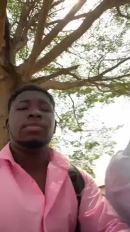 The video shows a dark-skinned man with short black hair wearing a light pink button-down shirt. He is looking at the camera, and a backpack is visible on his shoulders. Two other men are behind him, one wearing a white shirt. They seem to be sitting outside, as a large tree with many branches is visible behind them. The scene appears to be outdoors during daylight.
