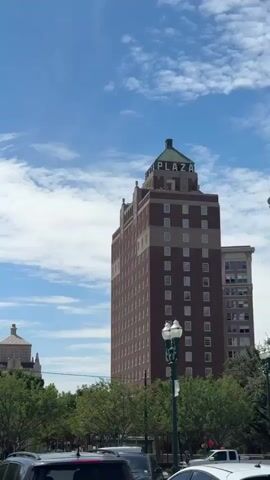 This is a vertical, outdoor, daytime video shot in what appears to be an urban environment. The video captures a variety of buildings, trees, and parked cars under a bright blue sky with scattered white clouds. On the right side of the frame, a tall, reddish-brown brick building with many windows dominates the view. A green-topped structure is visible at its apex, and the word "PLAY" is discernible on its side. In the middle ground, a smaller building with a distinctive domed roof and an intricate facade is partially obscured by lush green trees. To the left of this domed building, a tall lamppost with multiple light fixtures stands out. Further to the left, another building with a light-colored facade and numerous windows is visible. Closer to the foreground, a modern building features a prominent mural of a stylized animal, possibly a giraffe, rendered in vibrant, multi-colored mosaic tiles. Below the mural, a shipping container appears to be decorated with a colorful painting of a leopard wearing sunglasses. Several cars, including SUVs and sedans, are parked in the foreground, obscuring the lower portion of the buildings and trees. The video pans slowly from right to left, showcasing the different architectural styles and urban features. The overall impression is a snapshot of a bustling city center on a sunny day.