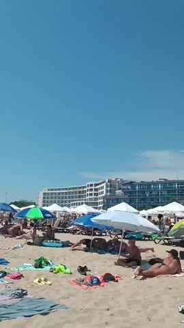 A crowded beach scene on a sunny day with a clear blue sky. Numerous people are scattered across the sand, many under umbrellas or beach canopies, relaxing on towels or lounge chairs. Some individuals are sunbathing, while others appear to be walking or interacting. In the background, a large, multi-story hotel or resort complex dominates the landscape, with a Ferris wheel and amusement park rides visible beyond it, suggesting a resort town. The beach is covered with towels, bags, and various beach accessories, indicating a popular recreational area.