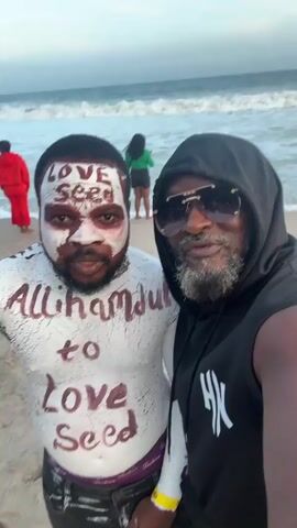 On a beach, two men stand close together, posing for a selfie. The man on the left is covered in white paint, with dark brown writing over the white, including "Love Seed" on his forehead and "Allhamdul To Love Seed" on his chest. The man on the right wears a black hoodie with a white logo on the front and sunglasses. A beach and ocean are visible in the background, with a few other people further away. The man on the left is pointing at the camera. Both men are smiling.