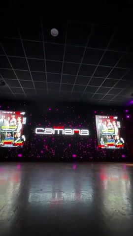 The video displays an indoor performance venue or club, characterized by a dark and modern aesthetic. The room is dimly lit, with the primary illumination emanating from three large LED screens positioned on the back wall. The floor is dark and highly reflective, mirroring the lights and images from the screens. The ceiling features a dark grid pattern with a single, round light fixture visible.

In the center of the back wall, a prominent white LED sign spells out "camana," with a small crown-like symbol above the first 'a'. This central display is surrounded by vibrant pink sparkling lights that animate across the screen. Flanking this central display are two larger LED screens, each showing what appears to be a promotional poster or advertisement.

On these side screens, the text "camana really" is visible at the top. Below this, "SAB 25" (likely indicating Saturday 25th) is displayed, along with the phrase "THE GREATEST SHOW." Each poster features an image of a group of people, possibly a band or performers, dressed in various styles, suggesting a musical or entertainment event.

Lively background music with vocals is playing, creating an atmosphere of anticipation or celebration within the empty venue. The setting suggests an event is either about to begin or has just concluded. The overall impression is that of a contemporary entertainment space in Buccinasco, Italy, ready for or hosting a "camana" event.