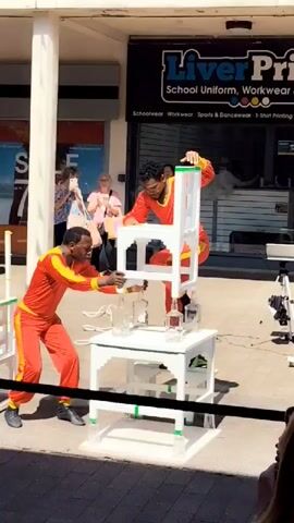 Two men in red and yellow athletic outfits are performing a balancing act outdoors. One man is on the ground, carefully manipulating several glass bottles that are positioned on a white table. The second man is standing on a chair, which is precariously balanced on top of the bottles. He is holding onto the back of the chair for support. In the background, there is a shop with a sign that reads "Liver Print School Uniform, Workwear". Several onlookers are visible, some of whom are taking pictures. The scene appears to be a public space, possibly a street or a pedestrian mall, with sunlight indicating daytime.