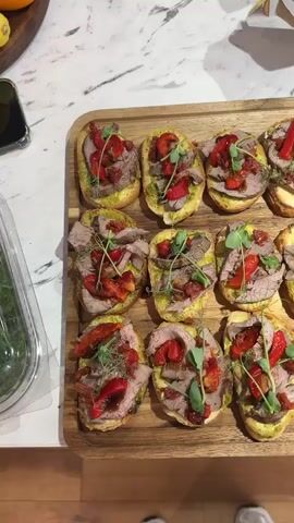 The video shows a top-down view of a kitchen counter with several platters of appetizers. The platters are arranged on a wooden cutting board. The first platter, on the left, contains approximately 12 pieces of bruschetta topped with sliced roast beef, roasted red peppers, and microgreens. The second platter, in the center, also on the wooden board, has about 10 pieces of bruschetta topped with fresh mozzarella, sliced tomatoes, sun-dried tomatoes, and microgreens, drizzled with balsamic glaze. The third platter, on the right, also on the wooden board, features approximately 10 pieces of bruschetta topped with shrimp, avocado puree, and microgreens. A person's feet are visible at the bottom of the frame, suggesting they are standing and filming the food. The kitchen environment appears to be modern with a marble countertop and stainless steel appliances visible in the background. The lighting is bright, highlighting the food.