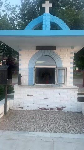 The video shows a small, white, brick building with blue trim and a cross on top. The building has an arched window with two small doors that are open. Inside the window, a statue of a religious figure can be seen, along with candles and other religious items. The building is located outdoors, with trees and a fence visible in the background. A person in a white shirt is briefly visible walking in the background. The overall scene appears to be a small chapel or shrine.