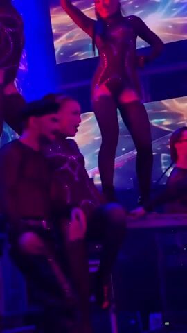 A dimly lit, vibrant performance space is captured. In the foreground, a woman in a black, sequined bodysuit with cutouts and thigh-high stockings dances suggestively. She is interacting with a man to her left, who wears a black sheer long-sleeved shirt and black leather pants, with a black beret on his head. He appears to be a performer as well, possibly a dancer or backup performer. Behind and above them, another woman in a similar black, sequined costume is positioned on a raised platform, her arms extended upwards. Red and blue stage lights illuminate the scene, creating a dramatic and energetic atmosphere. The background displays abstract, colorful lighting effects on a screen or projection. Other performers are visible in the periphery, also in dark, sparkling attire. The overall impression is of a lively and theatrical stage show.
