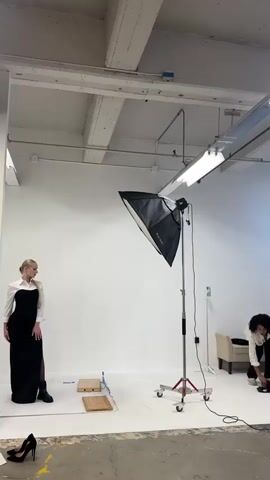 The video shows a fashion photoshoot taking place in a studio. A female model, wearing a long black dress with a white collar and boots, stands on a white backdrop. A large softbox light is positioned in front of her. A photographer, holding a camera, is on the right side of the frame, actively taking pictures of the model. In the background, there is a couch and a light-colored wall with exposed ceiling beams. On the floor in front of the model are two square props and a pair of black high-heeled shoes. The overall environment suggests a professional studio setting.