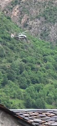 A white and black helicopter flies over a dense, green forest. The helicopter is tilted to the right and is moving from left to right across the frame. The background is a steep, rocky mountainside covered in trees. The sky is cloudy and overcast.