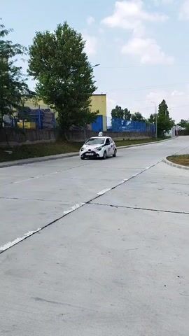 A white compact car with "Scoala de Soferi" (Driving School) markings on the side drives along a paved road. The car has a roof-mounted sign that says "L" indicating a learner driver. The road is in an industrial or commercial area with a concrete fence and buildings in the background. Trees and utility poles are also visible. The sky is partly cloudy. The car is moving away from the camera.