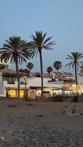 The video shows a beach scene at dusk. In the foreground, there is a sandy beach with pebbles. Two people are sitting on the sand, facing each other. In the background, there is a row of buildings with white walls and red-tiled roofs. Palm trees are scattered throughout the scene. The buildings appear to be restaurants or shops, with lights on and people visible inside and outside. The sky is a dusky blue. The overall atmosphere is peaceful and relaxed.