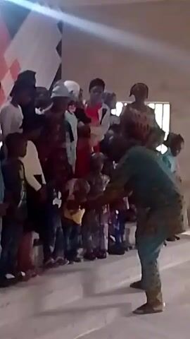 A medium shot depicts a group of approximately 15 people, predominantly children, standing on a raised platform indoors. The group is diverse in age and attire, with some wearing casual clothing like jeans and t-shirts, while others are in more traditional or formal wear. The environment appears to be a simple hall or gathering space, with a window visible and an unclear backdrop featuring black, white, and red colors. A man in a patterned green shirt and pants stands in front of the group, facing away from the camera, gesturing with his hands and appears to be leading or directing the group. The overall scene suggests a casual gathering or a simple performance, possibly in a community or religious setting.