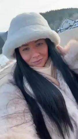 A woman in a fur coat and hat walks along a snowy street. She looks directly at the camera as she walks, and the camera is shaky as if she is holding it herself. The woman has long, dark hair, fair skin, and is wearing light makeup. Her coat is a light brown color and looks to be faux fur. Her hat is white and fluffy. The street is partially covered in snow, and there are houses and trees in the background. The sky is overcast.