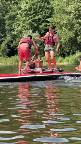 The media file captures a lively scene on a river in Warisy, Belgium, on a sunny day. The primary subjects are a group of people engaged in playful roughhousing on a large, red and white inflatable paddleboard. Two individuals are particularly active, one wearing red shorts and a life vest, and the other in dark shorts and a life vest. They are wrestling and playfully pushing each other, with one person attempting to push the other off the board. A third person, partially visible, is also on the paddleboard, seemingly reacting to the playful struggle. Further in the background, another person is visible on a separate inflatable object.

The setting is a tranquil river with calm, reflective water. Lush green trees and foliage line the riverbank, suggesting a warm, summer day. The sunlight indicates it's likely midday, casting bright light on the scene. The overall mood appears to be one of lighthearted fun and recreation.

Visible on the paddleboard is the text "TIKI FACTORY" printed in white. The people are dressed in casual summer wear, including life vests, which are essential safety gear for water activities. The dynamic poses and expressions of the individuals suggest an enjoyable and energetic interaction.