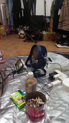 The video shows a close-up of a small, blue, portable fan with its blades spinning. The fan is positioned on a reflective, silver-colored surface, possibly a tarp or mat. In the foreground, to the left of the fan, a clear plastic bottle with a white cap is visible, and next to it, a package of tissues with a cartoon character on it. To the right of the fan, there are several white power adapters plugged into what appears to be a power strip. A partially filled ashtray with cigarette butts is in the lower center of the frame, to the left of the bottle. The background is blurred, but hints at a room with clothing hanging on a rack and a soccer ball on the floor. The lighting suggests an indoor environment.