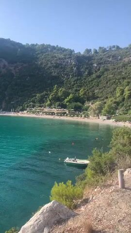 The video pans across a serene turquoise bay with a sandy beach nestled at the base of a lush, green, mountainous backdrop. The water is calm, with gentle ripples reflecting the clear blue sky. Several small buoys dot the water. A small white motorboat is anchored near the shore. A larger white sailboat is visible further out in the bay. The beach appears sparsely populated, with a few umbrellas and loungers set up. The overall impression is one of tranquility and natural beauty.