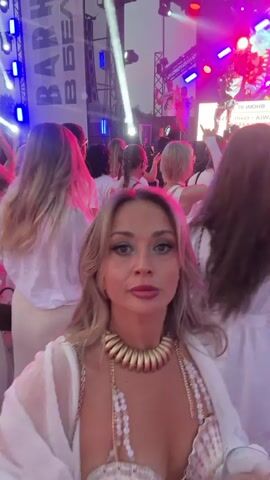 The video shows a group of people at an outdoor event, possibly a concert or festival, at night. The lighting is predominantly blue and red, with stage lights visible in the background. In the foreground, a woman with blonde hair, wearing a white dress and a shell-themed bikini top, turns to the camera and smiles. She is holding a drink. To her right, a woman with dark hair and bangs, also in a white dress, is seen interacting with the blonde woman. Other people in white attire are visible in the background, dancing or socializing. A large banner with Cyrillic text is also present. The overall atmosphere appears festive and energetic.
