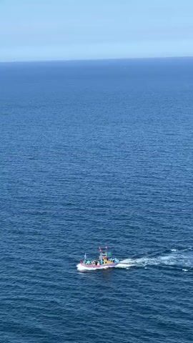 A small, multi-colored fishing boat with a white hull and a red roof is sailing across a vast, deep blue ocean. The boat is moving from left to right, leaving a white wake behind it. The ocean is calm with small ripples on the surface. The sky is a light blue, with a faint haze on the horizon. In the far distance, two other very small white boats or buoys are visible. The shot is taken from a high angle, looking down on the boat and the water.