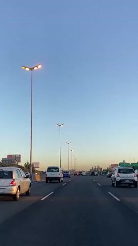 The camera shows a highway view from inside a car. Multiple lanes stretch out with traffic moving forward under a clear blue sky with a tinge of orange at the horizon. Streetlights illuminate the sides of the highway. Billboards and green vegetation dot the landscape on either side of the road.