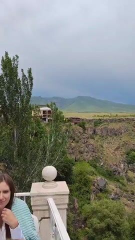 The video shows a wide view of a scenic landscape with a deep canyon. In the foreground, a white railing and a decorative sphere are visible, suggesting a viewing platform or balcony. A young woman is partially visible in the lower left corner, looking towards the camera. Beyond the railing, the canyon opens up, revealing steep, rocky walls covered in vegetation. A small waterfall can be seen cascading down one of the canyon walls. In the distance, rolling green hills and mountains form the backdrop under a cloudy sky. Some man-made structures, possibly buildings or ruins, are scattered along the canyon rim and on the distant hills. The overall impression is one of natural beauty with some signs of human presence.