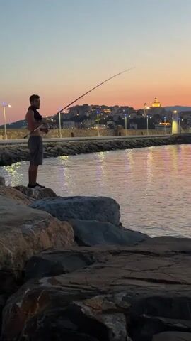 A young man stands on large rocks by the water, fishing with a rod. He is shirtless, wearing grey shorts, and black shoes. The water reflects the lights from a town in the background, which is situated on a hill. The sky is a gradient of orange and blue, indicating it is either sunrise or sunset. A lighthouse is visible in the distance. The scene appears to be a peaceful evening at a harbor or bay. No text is visible in the media.