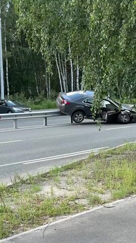 A dark gray sedan is heavily damaged and lodged on top of a guardrail on the side of a two-lane road. The front end of the car is crumpled, and the driver's side door is open. The rear of the car is tilted upwards, with the rear wheels off the ground. Trees and foliage surround the scene, with a forest visible in the background. Another car, possibly a dark-colored sedan, is visible further down the road, stopped and facing the same direction. The road appears to be asphalt, with white lane markings. The weather appears to be overcast or shaded, suggesting it is daytime.