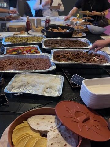 The video opens with a close-up shot of a buffet table featuring a variety of dishes served in aluminum trays. Visible foods include what appears to be corn, bell peppers and onions, two trays of brown beans, two trays of cooked meat (one specifically labeled "carne asada" on a small chalkboard sign), and white rice. Bottles of condiments like ketchup, stacks of plates and cups, and a container of tortillas are also present. Several hands are seen reaching for food, indicating active serving. The camera then blurs, showing people gathered around the table, suggesting a social event or meal service. Following this, the video focuses on a woman with long, dark, curly hair, wearing a black leather jacket over a white top, and a nose piercing. She is seen looking downward. The final shots are blurred as the camera moves, showing glimpses of an indoor setting with tables, chairs, and other people in the background, possibly an event space or studio. The event takes place in Culver City, United States of America.
