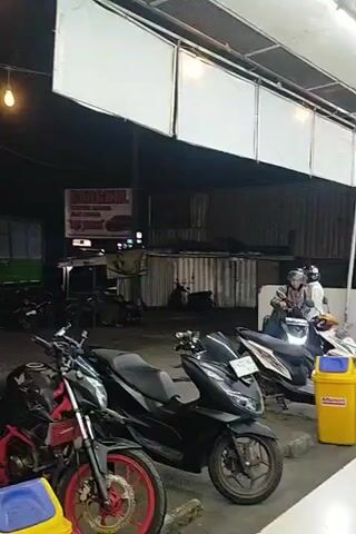 The media file captures an outdoor scene at what appears to be a roadside establishment, possibly a workshop or a food stall, in Lungbenda, Indonesia. The time of day is evening or night, as indicated by the artificial lighting from bulbs strung overhead and the general darkness of the background.

In the foreground, several motorcycles are parked. Two are particularly prominent: a black sportbike with red accents on its wheels and a black scooter. A red scooter with a green helmet on it is also visible. In front of the red scooter is a yellow and blue trash bin. Further back, more motorcycles are parked, and a few people are gathered, some appearing to be working or socializing.

Dominating the background is a large green truck with a silver cargo container. Behind the truck and the motorcycles, there are structures that suggest a commercial or service area. A sign is visible hanging from a pole, with text that reads "BENGKEL" (which translates to "workshop" in Indonesian), followed by "MOTOR", "CAT DUCO", and "LAS JUNI JAYA". This confirms the presence of a workshop offering services like motorcycle repair, paint (duco), and welding.

Two individuals are standing near a scooter, one of them appears to be wearing a helmet and is facing away from the camera. Another person, a man in a black t-shirt, is walking into the frame from the right. The overall atmosphere seems casual and functional, typical of a local business operating in the evening. There are no obvious indications of specific weather conditions, but the lighting suggests it is likely not raining.