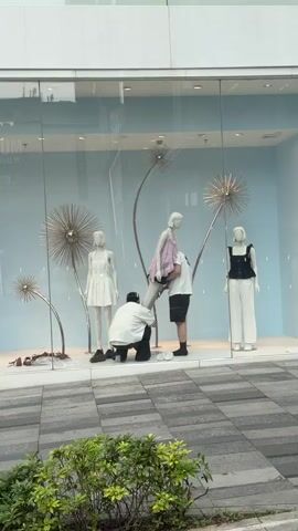 The video shows a store window display with three mannequins and two people interacting with them. One mannequin on the left wears a white dress. A second mannequin in the center wears a pink and white patterned dress. A third mannequin on the right wears a black top and white pants. Two individuals are present, both appearing to be male. One person, wearing a white shirt and dark pants, kneels in front of the center mannequin, seemingly adjusting its leg. Another person, wearing a white t-shirt and black shorts, stands behind the center mannequin, also appearing to be adjusting it. The background of the store window is light blue with decorative metallic dandelion-like structures. The floor outside the window is paved with gray and black tiles, with some green foliage visible in the foreground.