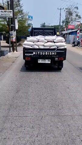 The video shows the rear view of a black Mitsubishi pickup truck driving on an asphalt road. The truck bed is loaded with multiple white sacks of material, tied down with ropes. The license plate of the truck reads "A 8505 BN". A motorcycle with a rider wearing a white helmet is passing the truck on the right side. The environment appears to be a rural or suburban area with trees and buildings visible on the sides of the road. The sky is clear and the weather seems to be sunny. The camera is positioned behind the truck, following its movement.