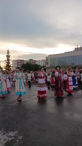A group of people in traditional clothing are dancing in a circle outdoors. The people in the foreground are women wearing long, brightly colored dresses and flower wreaths in their hair. They are holding hands and moving in a synchronized dance. In the background, there are more people, some of whom appear to be watching the dancers. There are buildings and trees in the distance, and the sky is overcast. The ground is wet, suggesting that it may have recently rained.