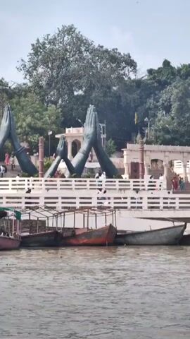 The video shows a wide shot of a body of water with several boats docked along the shore. In the background, there are large statues of hands in a prayer position, with stairs leading up to a building. Trees and people are visible in the background, and the sky is overcast. The water appears murky and brown, and the boats are of various sizes and colors, some with canopies.