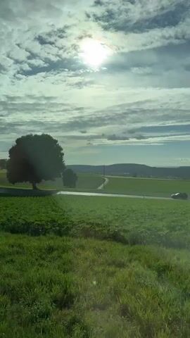 The camera captures a rural landscape through a vehicle window, looking at green fields, a road, a tree, and distant hills. The sun is high in the sky, and there are scattered clouds. A dark-colored truck with a white trailer, labeled "PLANZER," drives along the road. A white car follows the truck, and a dark car is seen further down the road in the opposite direction. A white and black road marker is visible in the foreground. The sky changes from partially cloudy to mostly clear blue.
