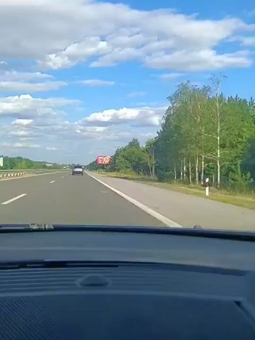 The video, captured from inside a vehicle, shows a highway scene under a partly cloudy sky. The perspective is from the driver's seat, looking forward through the windshield. A lane divider line marks the right side of the road, separating it from a shoulder. Beyond the shoulder, a line of trees, including birch trees, forms a natural barrier. In the distance, a billboard with a KFC advertisement is visible on the right side of the highway. The billboard features an image of fried chicken and text indicating the distance to KFC as "2 km." Another car is seen driving in the same direction on the highway, further ahead. The overall environment suggests a road trip or travel scenario on a clear, sunny day.