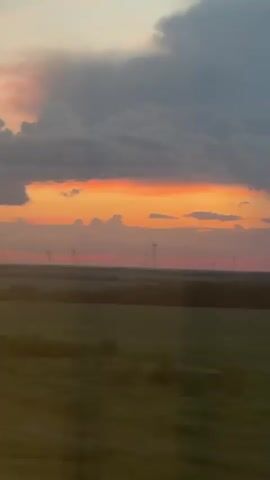 A video shows a sunset over a field with wind turbines. The sky is filled with colorful clouds ranging from dark gray to bright orange. A strip of orange is visible across the horizon, with lighter shades of pink and purple above and below it. In the distance, several wind turbines are silhouetted against the sky. The foreground shows a dark field, possibly farmland, with some blurry details suggesting movement. The overall impression is of a peaceful, rural landscape at dusk.