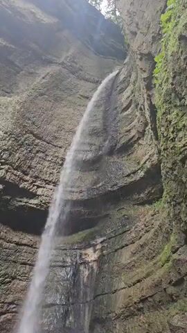 A waterfall cascades down a sheer rock face within a narrow canyon. The rock is textured and layered, with patches of moss and vegetation clinging to its surface, particularly on the right side. The water falls in a continuous stream, appearing frothy at its base where it meets the rocky ground. The upper portion of the canyon walls is visible, suggesting a dense canopy of trees overhead. The lighting is diffused, indicating an overcast day or a shaded environment.