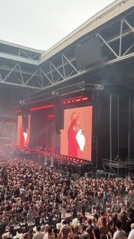 The video shows a wide shot of a large outdoor concert stadium filled with a massive crowd of people. The stage is at the far end of the stadium, featuring two large screens displaying a performer. The performer is a young man wearing a red shirt, holding a microphone and a white cloth. The screens are illuminated with a bright orange-red hue, and there are several rows of red lights above the stage.

The crowd is densely packed, with many people standing and cheering. The overall atmosphere appears energetic and celebratory. The shot is taken from a higher vantage point within the stadium, looking down towards the stage and the audience. The stadium itself is a modern structure with a visible roof and seating areas. The sky is visible above the stadium, suggesting it is daytime. There is no text clearly visible in the media. The scene captures the excitement of a live musical performance at a large venue.