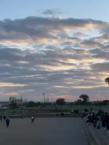 The video shows a soccer field with a group of people standing on a low wall overlooking the field. A player in a neon yellow jersey is visible on the field. The sky is filled with a mix of gray and white clouds, with patches of orange and yellow light visible near the horizon, indicating either sunrise or sunset. A silhouetted tree stands near the edge of the field, its branches partially obscuring the sky. The scene suggests an outdoor gathering, possibly watching a soccer practice or game.
