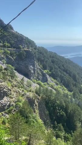 The video shows a scenic view from a mountain overlooking a body of water with islands. The mountain slope is covered in lush green trees and rocky terrain. The sky is clear and blue with the sun shining. There are no people or specific objects clearly visible in the video, except for what appears to be a faint line or cable in the upper left corner of the frame. The camera appears to be in motion, slowly panning across the landscape.