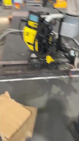 A yellow and black welding machine on wheels moves along a metal track on a concrete floor. A person in a red shirt and blue pants, wearing gloves, is operating the machine, which appears to be cutting or welding metal. The environment looks like a workshop or factory, with various industrial equipment visible in the background, including a forklift. The machine has a digital display and several hoses and cables connected to it. The metal being worked on is a long, flat piece.