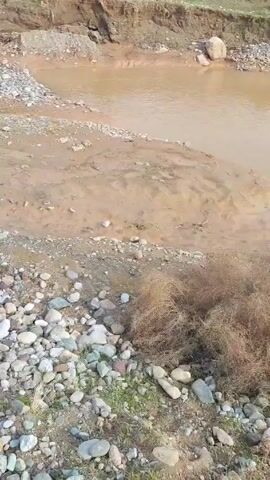 The video depicts a rocky, muddy landscape, likely a riverbed or floodplain with several small pools of muddy water. The ground is covered in a mix of pebbles, small rocks, and some sparse vegetation. In the distance, a range of snow-capped mountains is visible under a cloudy sky. There are some utility poles in the mid-ground and hints of buildings and structures, possibly a small village or town, also in the mid-ground. The overall impression is one of a barren, slightly desolate landscape, with signs of erosion and water flow.