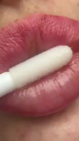 The video shows a close-up of a person's lips. They are applying a clear lip gloss with a white applicator wand. The lips are a natural pink color with visible texture and some fine lines. The skin surrounding the lips is fair with a few small spots or blemishes. The lighting is bright and even.