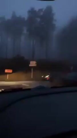 The video, shot from the dashboard of a car, shows a dark, foggy scene at a road intersection. There are road signs pointing to different destinations. Tall trees line the roadside, obscured by thick fog. A car with its headlights on is visible at the intersection, driving away from the camera. The overall ambiance is dark and slightly eerie due to the fog and low light.