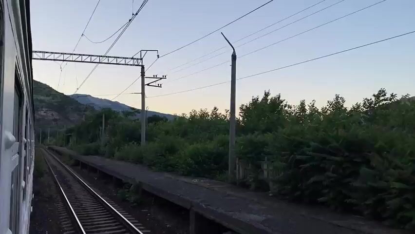 The video shows a view from a train window, looking out at a train station platform. The platform is empty and appears to be in a rural area. There are trees and bushes lining the side of the platform, and mountains can be seen in the distance. The sky is clear and it appears to be dusk. The train tracks are visible in the foreground, and the train is moving along them. There are overhead power lines for the train.