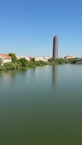 A wide, still river fills the foreground of the video, its surface a deep green. On the left bank, lush trees and vegetation are visible, interspersed with a few buildings. Further inland, a very tall, slender, curved skyscraper dominates the skyline. It is a reddish-brown color and casts a distinct reflection on the water. To the right of the skyscraper, more buildings are visible, as well as a bridge spanning the river in the distance. The sky is a clear, bright blue. Two small figures in a boat are visible on the river on the far left side of the frame.