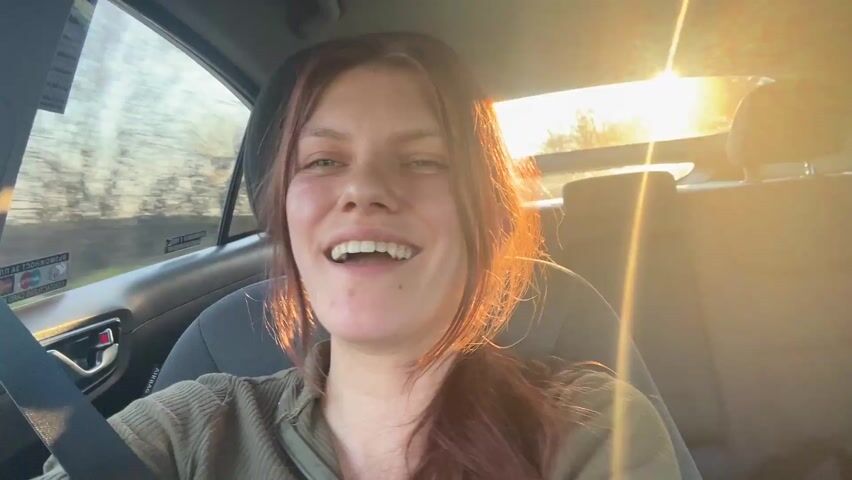 The video shows a young woman in the driver's seat of a car. She is wearing a green shirt and a seatbelt. Her hair is a mix of brown and red, and she is smiling and laughing. The sun is shining brightly in the background, creating a lens flare effect. The car appears to be moving on a road surrounded by trees. At one point, another arm reaches into the frame and playfully touches her face. Loud music is playing in the background.
