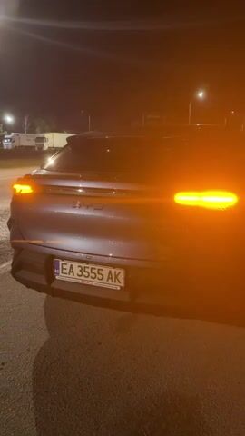 The video shows the rear of a dark-colored BYD vehicle parked at night. The vehicle's license plate, EA 3555 AK, is visible. The right turn signal flashes on and off. In the background, there are other vehicles, including trucks, and streetlights illuminate the area.
