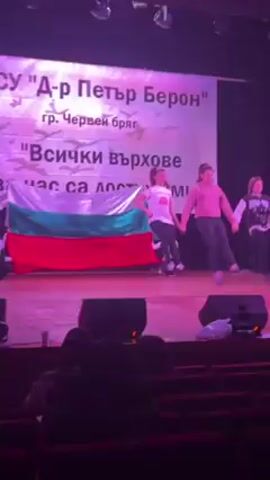 The video shows three young women holding hands and dancing on a stage, with a Bulgarian flag and a sign with Cyrillic text in the background. The sign refers to a school named "Dr. Petar Beron" in Cherven Bryag. The text on the sign says “All peaks are attainable for us!". They are wearing casual clothing. The stage is lit with purple light. There are some audio speakers in front of the stage, and the audience is sitting in red seats. There is a person recording the video.