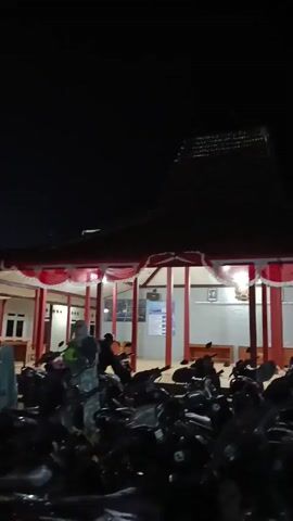 A vertical video shows a dark outdoor scene at night. In the foreground, a row of parked motorcycles is visible. Behind the motorcycles is a building with a distinct tiered roof. Red and white fabric decorations are draped along the front of the building. Several people are present, some of whom are interacting with each other or moving around the building's entrance. The lighting is dim, suggesting artificial illumination within and around the building. The sky is completely dark.